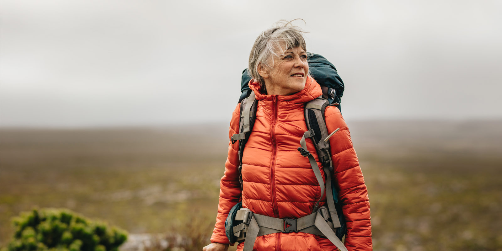 Ältere Frau mit grauem Haar und roter Wanderjacke blickt lächelnd in die Ferne in einer weiten Naturlandschaft.