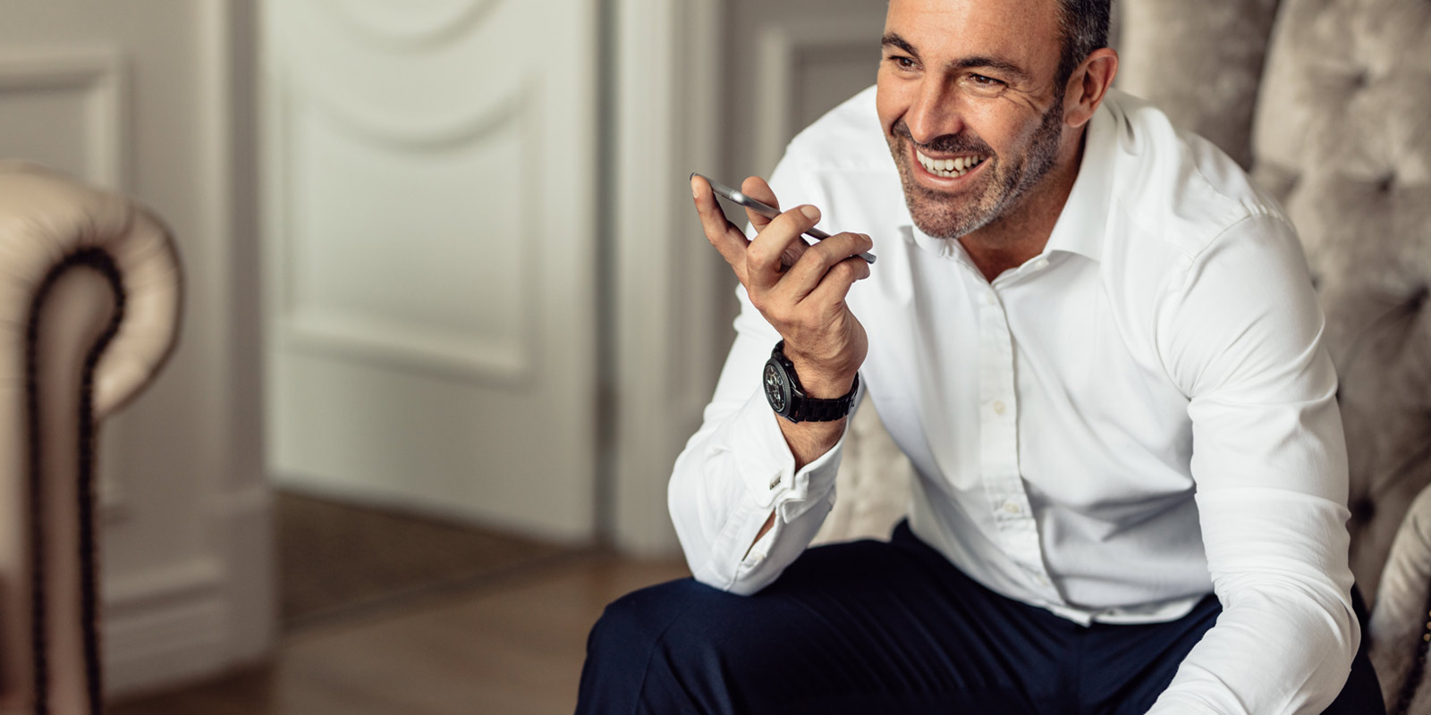 Mann in weißem Hemd spricht lachend in ein Smartphone, sitzt auf einem Sofa.