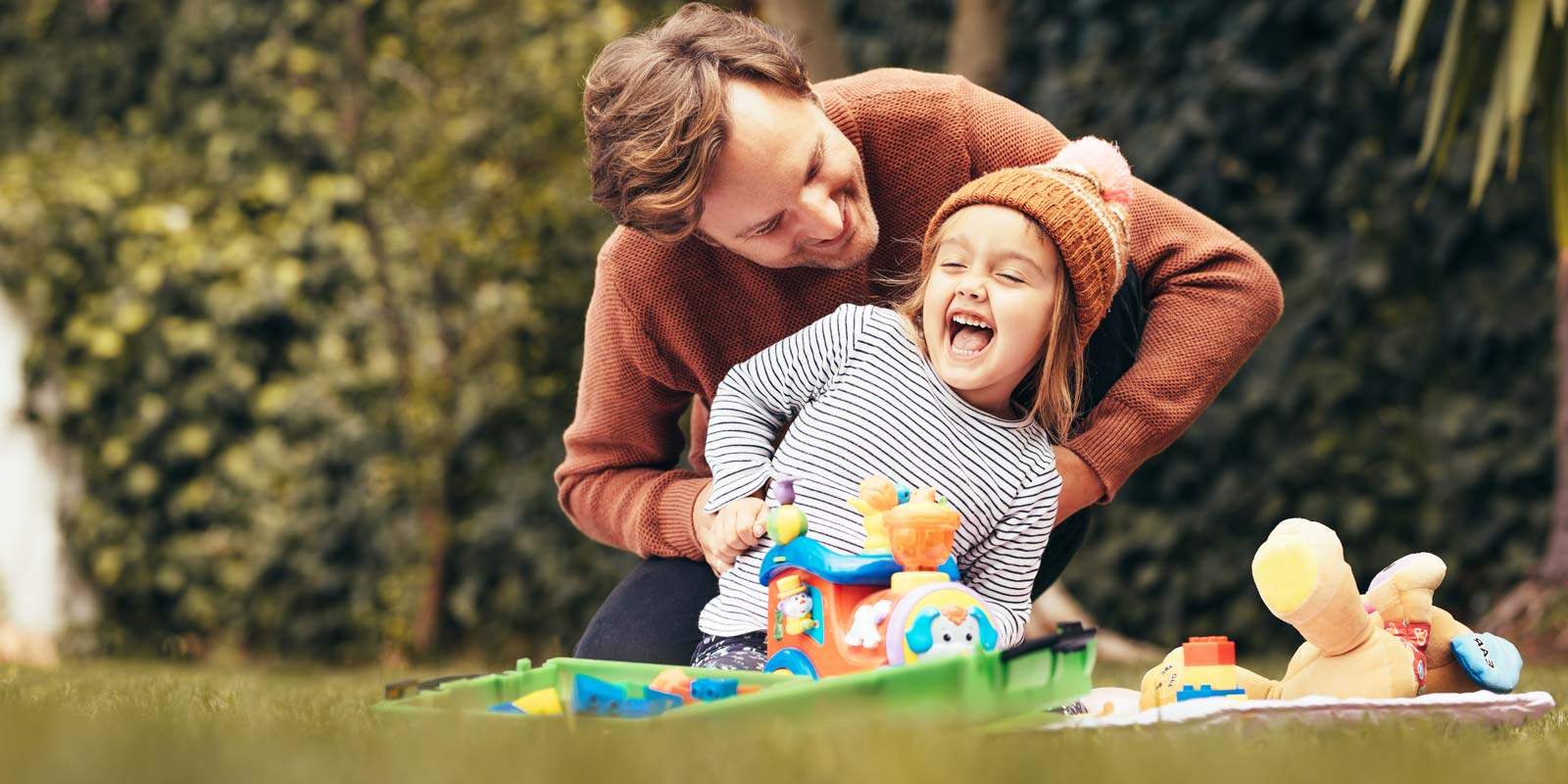 Vater spielt mit seiner kleinen Tochter lachend im Garten, umgeben von buntem Spielzeug.