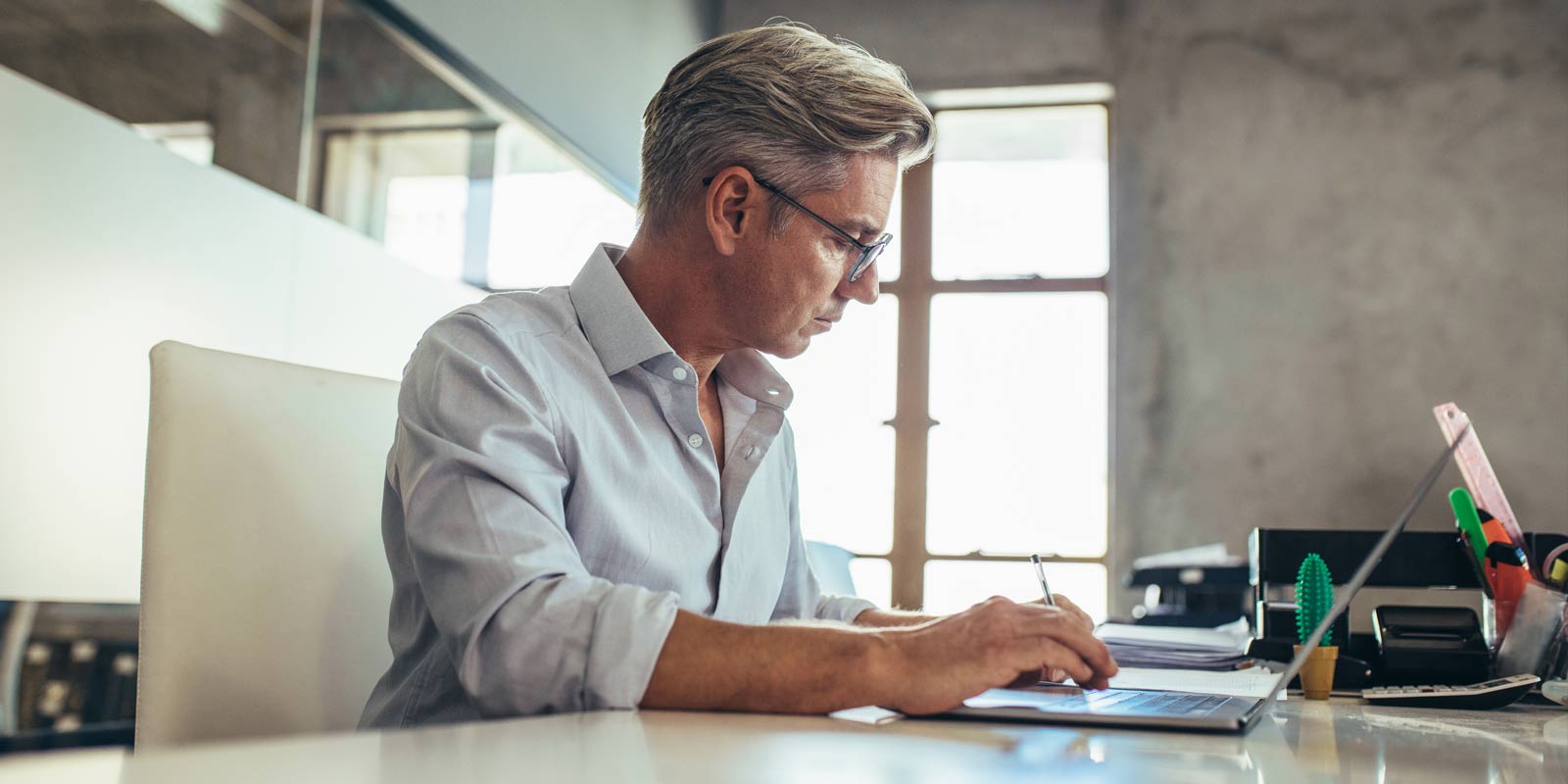 Geschäftsmann mittleren Alters mit grauen Haaren und Brille sitzt an einem hellen Schreibtisch, tippt auf seinem Laptop und hält einen Stift – konzentrierte Büroarbeit in modernem Arbeitsumfeld.
